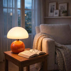 Nordic Ceramic Mushroom Table Lamp with orange ceramic base and white dome shade on a wooden side table beside a sofa.