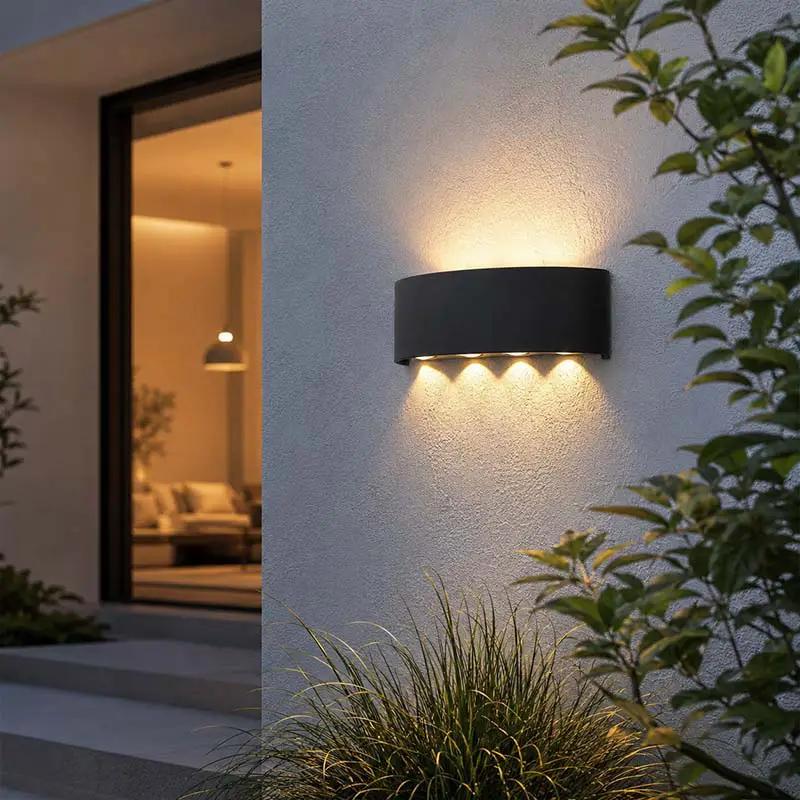 A modern black cylindrical outdoor wall light mounted on a textured white stucco wall, casting a warm yellow glow downward and upward. The scene is set in a real environment with green plants in the foreground and a warmly lit interior visible through a glass door in the background.
