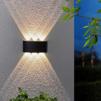 A modern black rectangular outdoor wall sconce mounted on a textured stucco wall, casting a warm light upward and downward in a V-shape. A split-screen view on the right shows the same fixture in use at dusk against a dark blue sky.