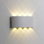 A modern rectangular wall sconce light fixture mounted on a white wall, casting warm light upward and downward in a diamond pattern.