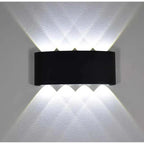 A modern black wall-mounted light fixture with three upward-facing and three downward-facing illuminated beams, shown against a textured white wall.