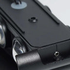 Close-up of a black metal camera accessory or mount with screws, ridges, and part of a lens visible, photographed on a light neutral background.
