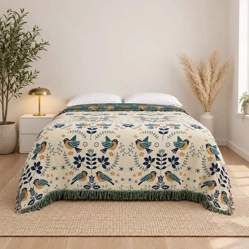 A patterned woven blanket with a fringe edge is styled on a bed in a bright, modern bedroom. The blanket features a cream background with a repeating pattern of blue birds, green foliage, and small orange floral accents.