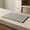 A rectangular gray slatted mat or tray with rounded corners, placed on a light beige countertop in a modern kitchen setting with wood cabinetry visible in the background.