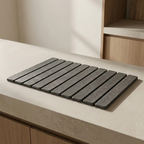 A rectangular dark gray slatted mat or trivet placed on a light beige countertop in a kitchen setting with wood cabinetry visible in the background.
