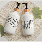 Two white soap dispensers labeled 'Dishes' and 'Hands' on a woven mat with leaves.